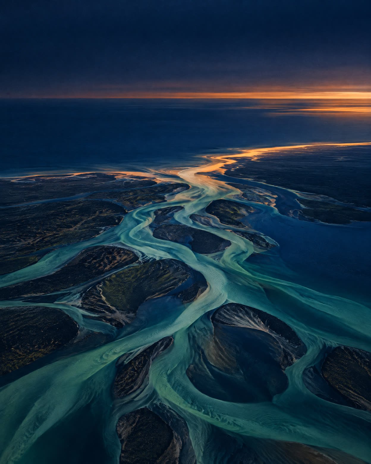 Abstract aerial composition of jade and indigo waterways meeting a deep blue sea at dusk.